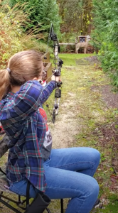 A woman draws a bow and arrow to practice shooting from a seated position.