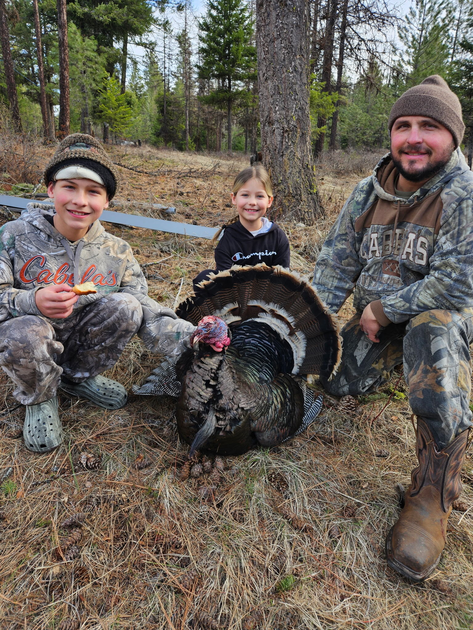A man and two children, dressed in camouflage, pose with a harvested turkey in a wooded area