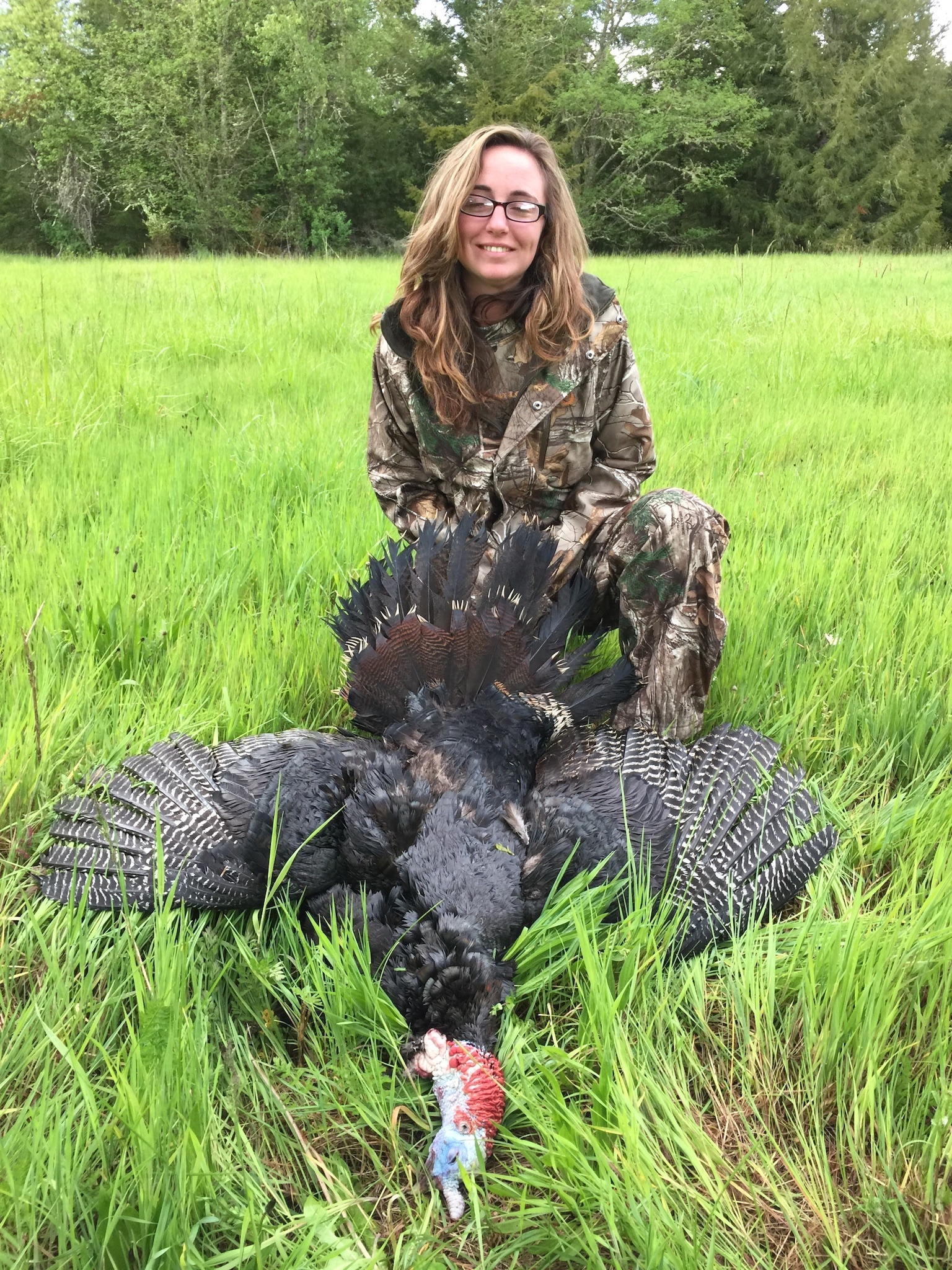 A young women in glasses and dressed in camouflage poses om tall green grass with her harvested turkey.