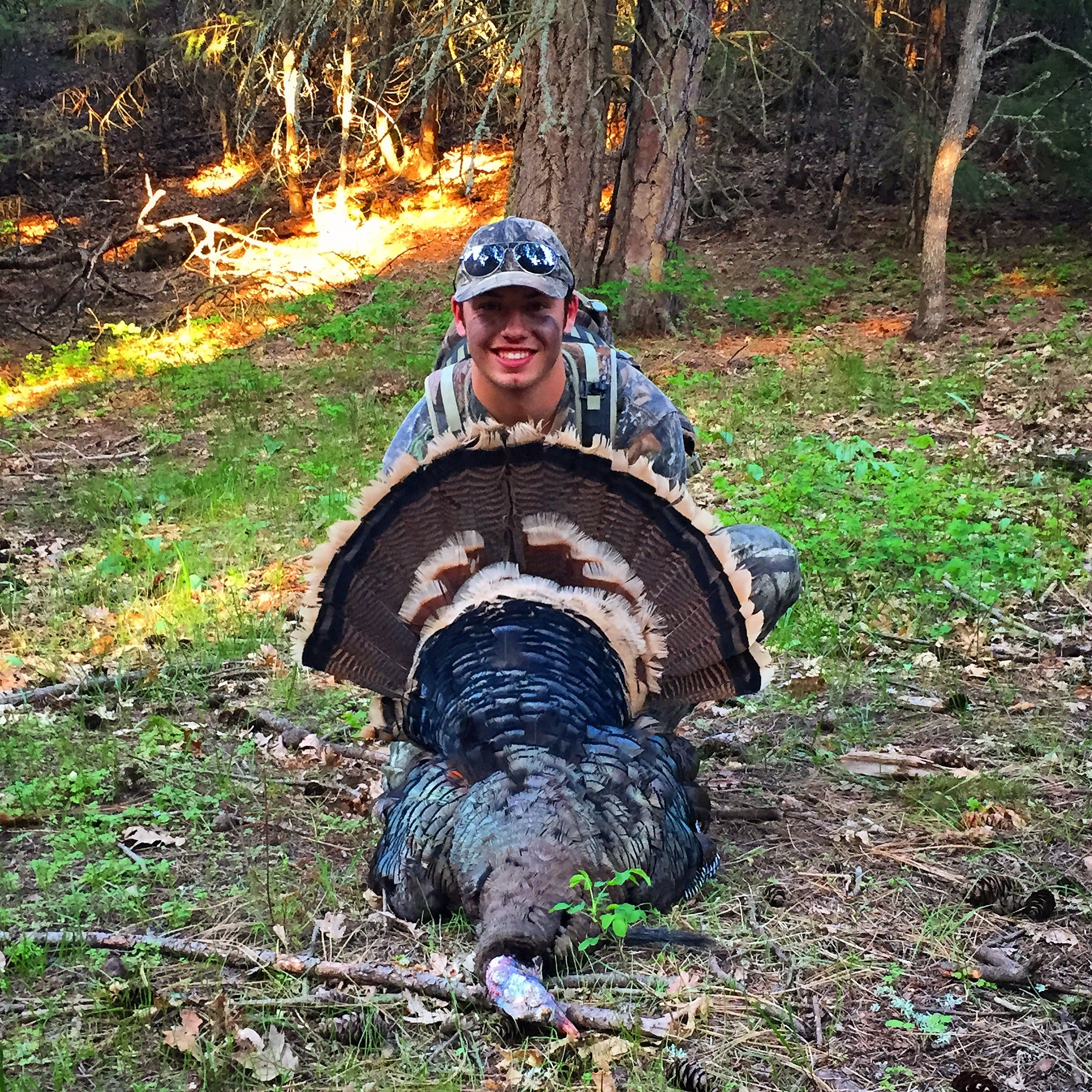 A smiling young man kneels on the ground while holding his harvested turkey.
